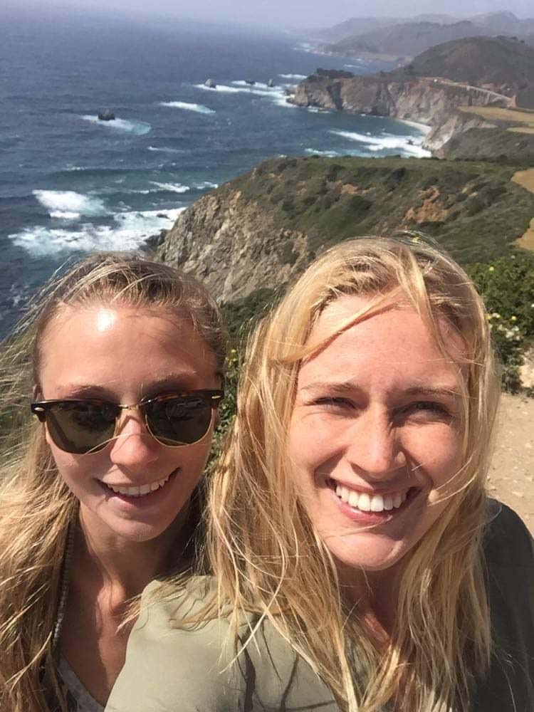 Två unga tjejer tar en selfie vid en bergsklippa med utsikt över havet